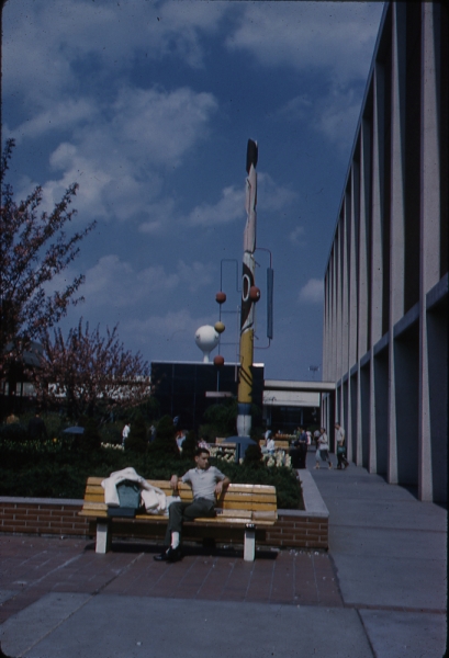 View of sculpture of totem pole by Gwen Lux at Northland Mall in Southfield, Michigan. Man sits on bench in foreground. 5-13-61